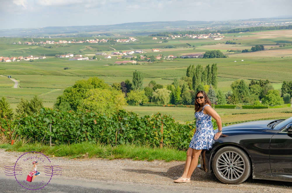 Harpreet in a blue dress leaning on a car in the vineyards